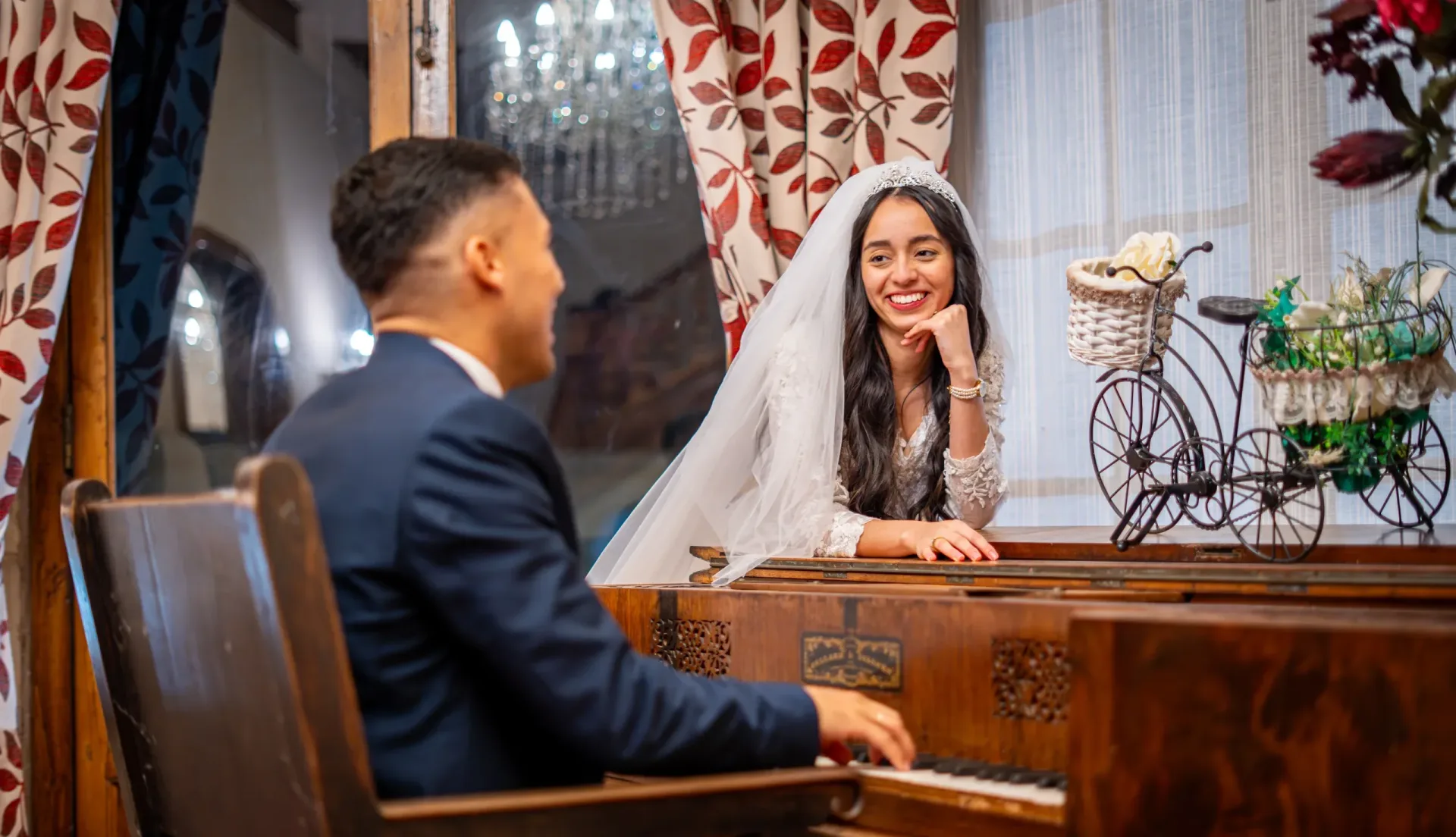 Novio tocando el piano para su novia en el día de su boda, ambos compartiendo una mirada romántica en un interior decorado.